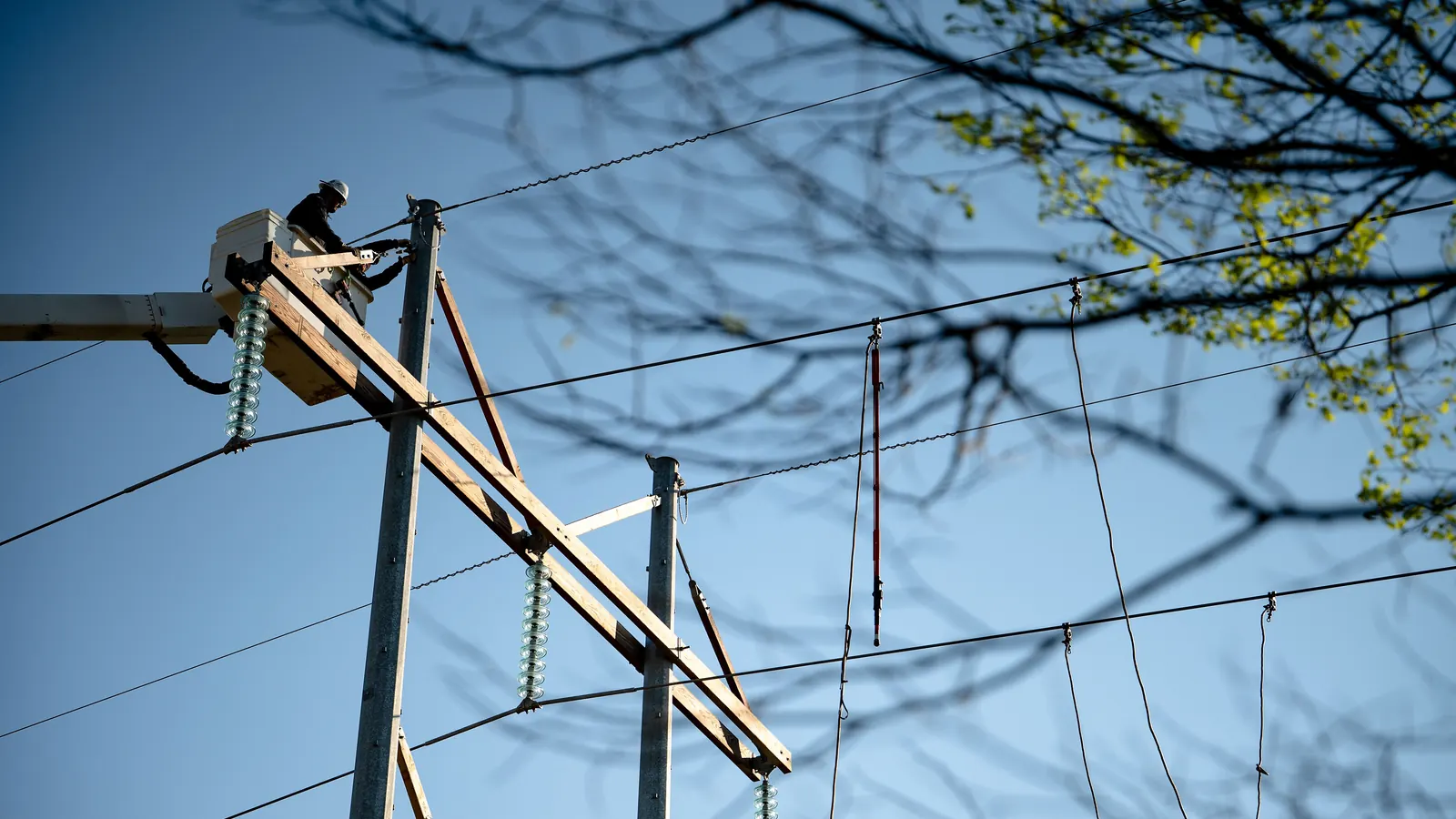 A worker in a hard hat in a white boom bucket works at a pole top with glass insulators, under a clear blue sky.