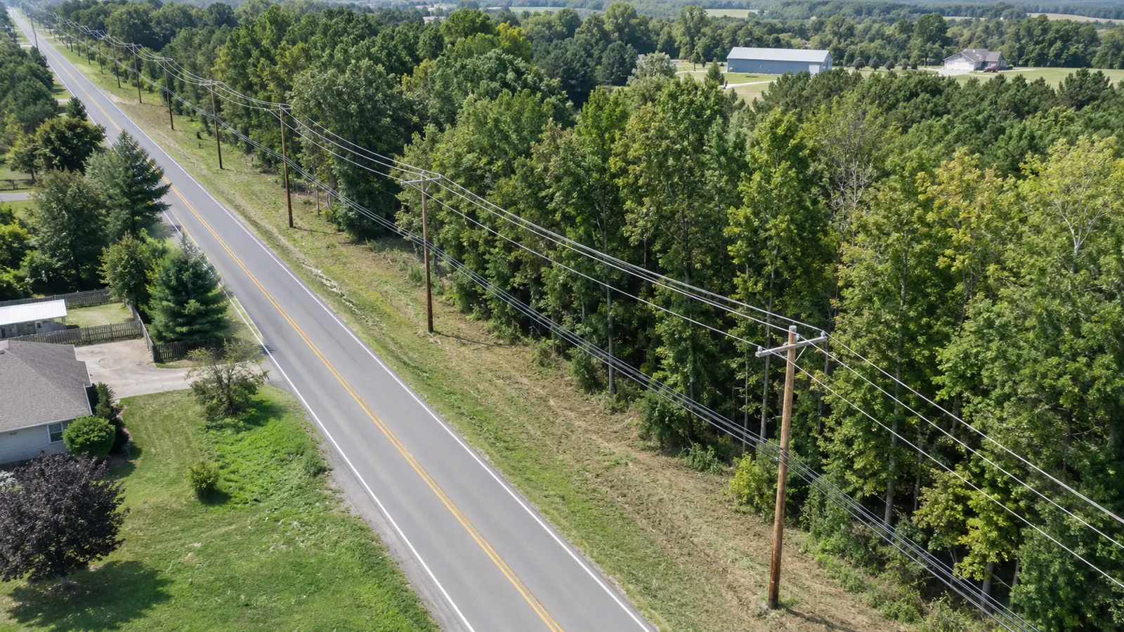 Oblique aerial view of a roadside utility corridor separated from trimmed tree canopy and nearby structures.