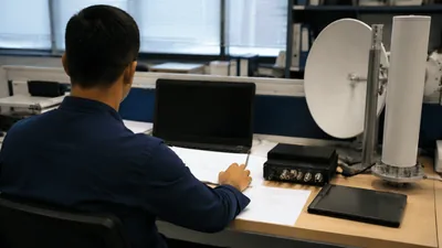 Engineer at an office desk writing on paper beside a laptop, with a dish antenna and radio equipment nearby.
