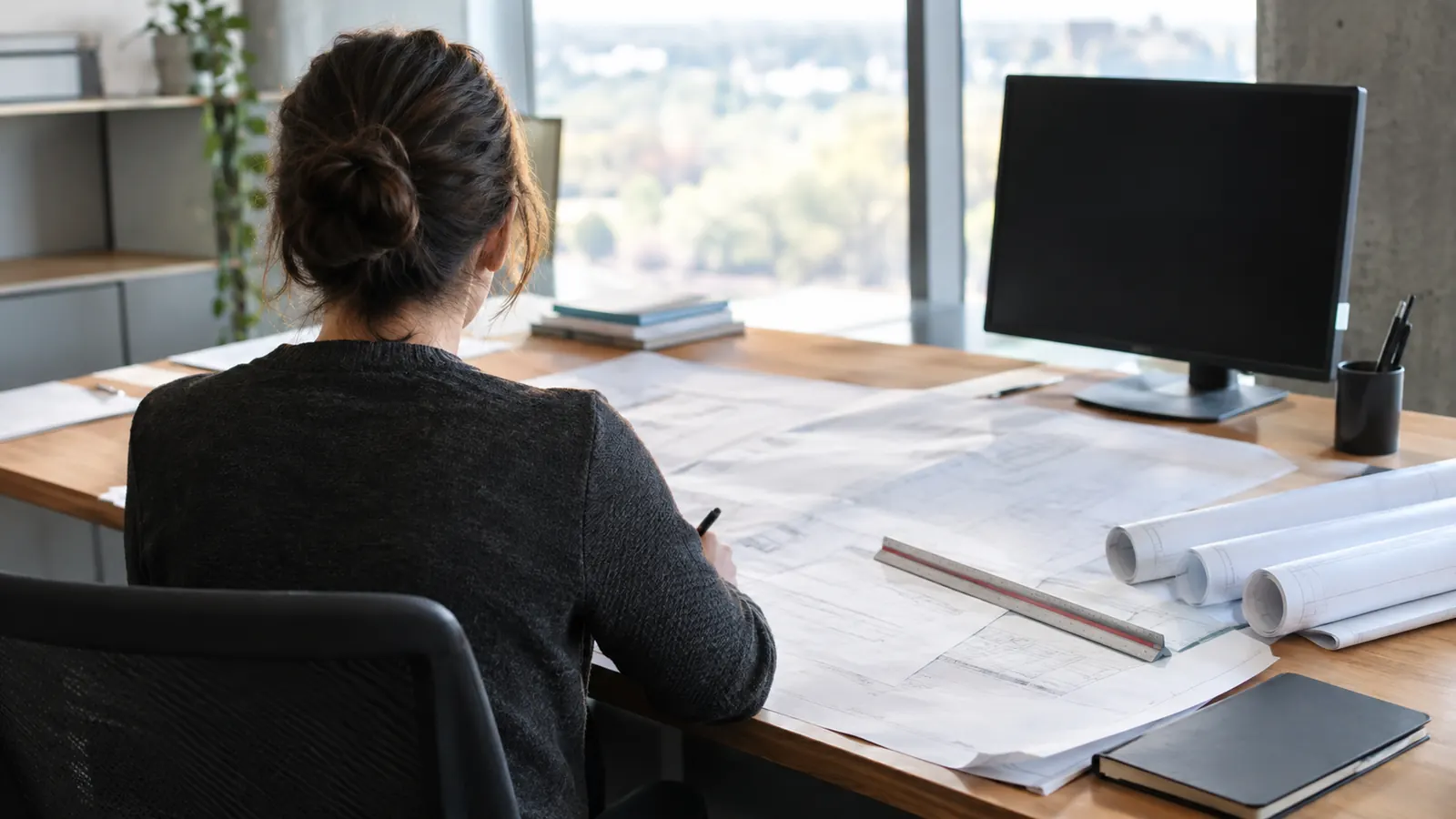 A person seated at an office desk by a window marks up spread-out drawings beside a scale ruler and a dark monitor.