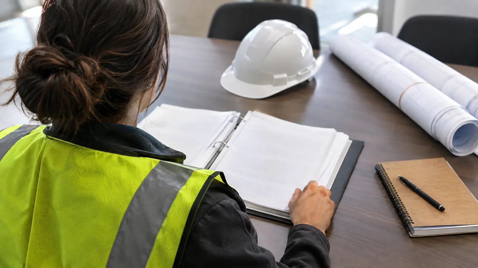 A person in a yellow safety vest reads a binder at a conference table beside a white hard hat and rolled plans.