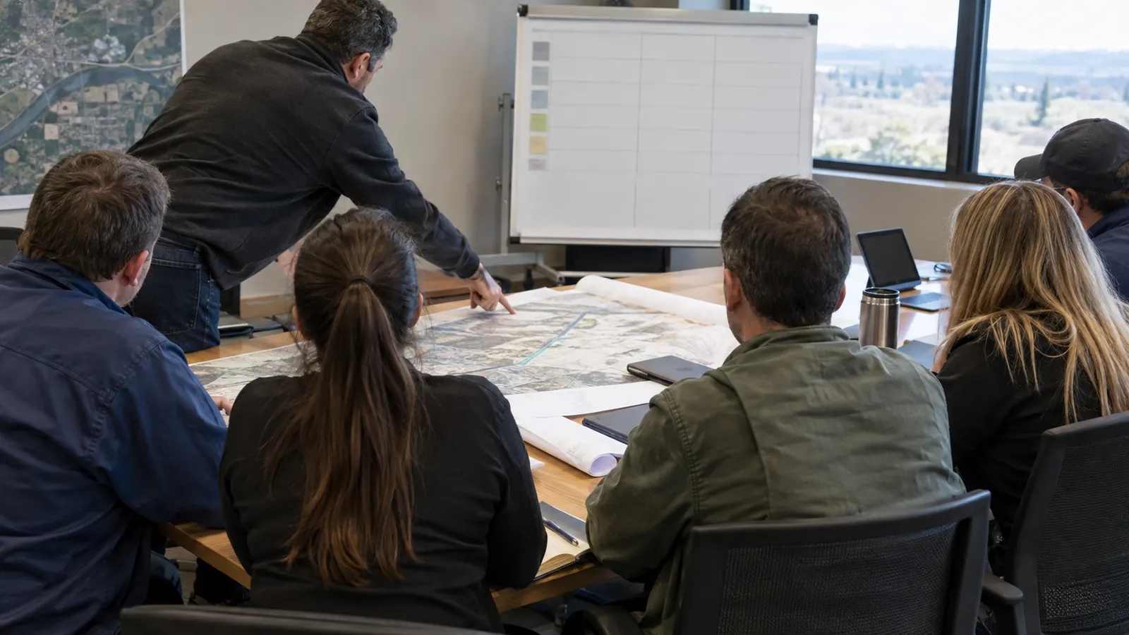 A man leans over a conference table pointing at a large aerial map as five seated people watch in an office.