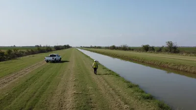 A worker in a yellow vest and white hard hat walks away along a grassy canal bank past a parked white pickup.