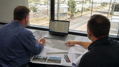 Permit coordinator reviewing unlabeled fiber route sheets beside rolled utility maps at a field office desk