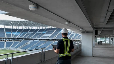 Technician using a handheld device beneath ceiling antennas in an empty stadium concourse