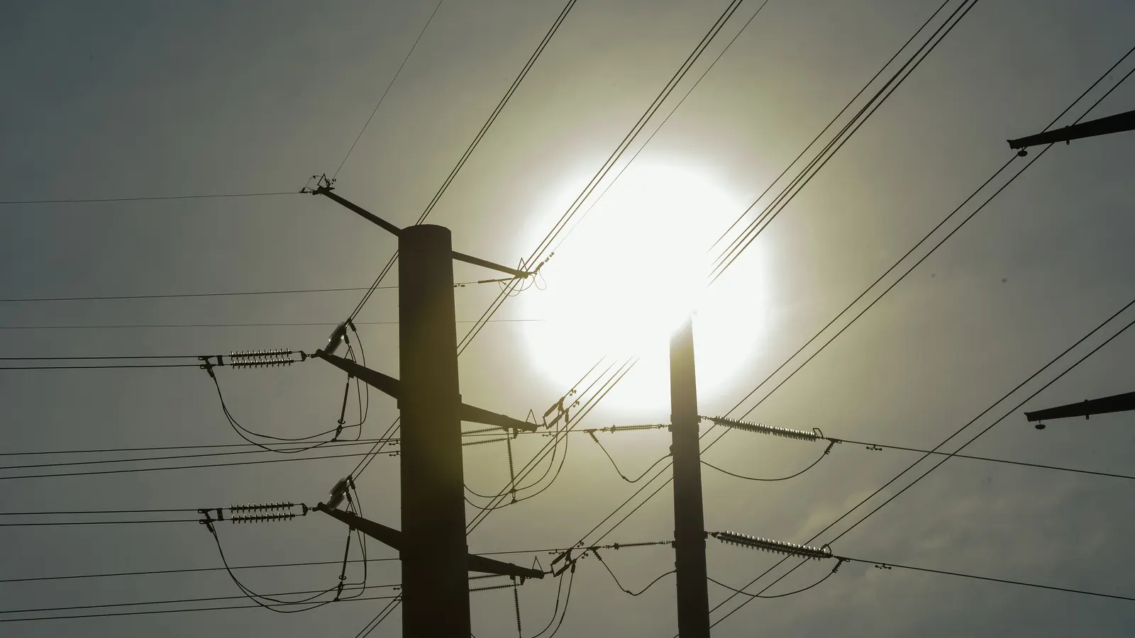 Two tall utility poles carry high-voltage power lines and strings of insulators, shown as dark silhouettes against a hazy sky with the sun glowing behind them.