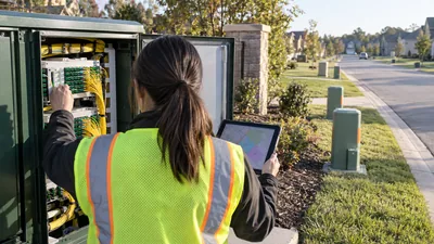 A worker in a safety vest holds a map tablet while reaching into an open green fiber cabinet on a suburban street.