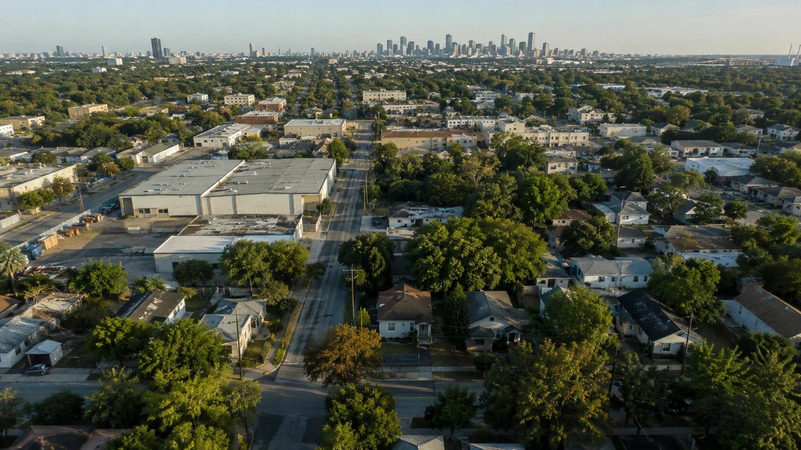 Oblique aerial view of a residential and light-industrial street grid leading toward a distant city skyline