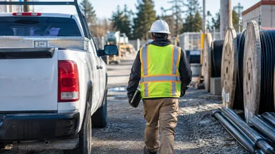 A worker in a hard hat and safety vest walks through a gravel yard past a white pickup truck and large cable reels.