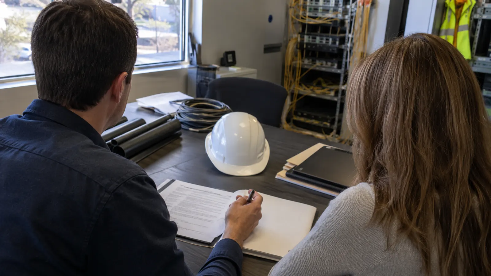 Two people at an office table look over papers as one holds a pen, with a white hard hat and cabling racks nearby.