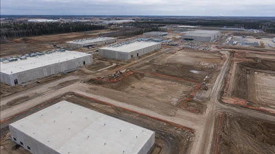 Oblique aerial view of concrete data center buildings and graded construction corridors under an overcast sky
