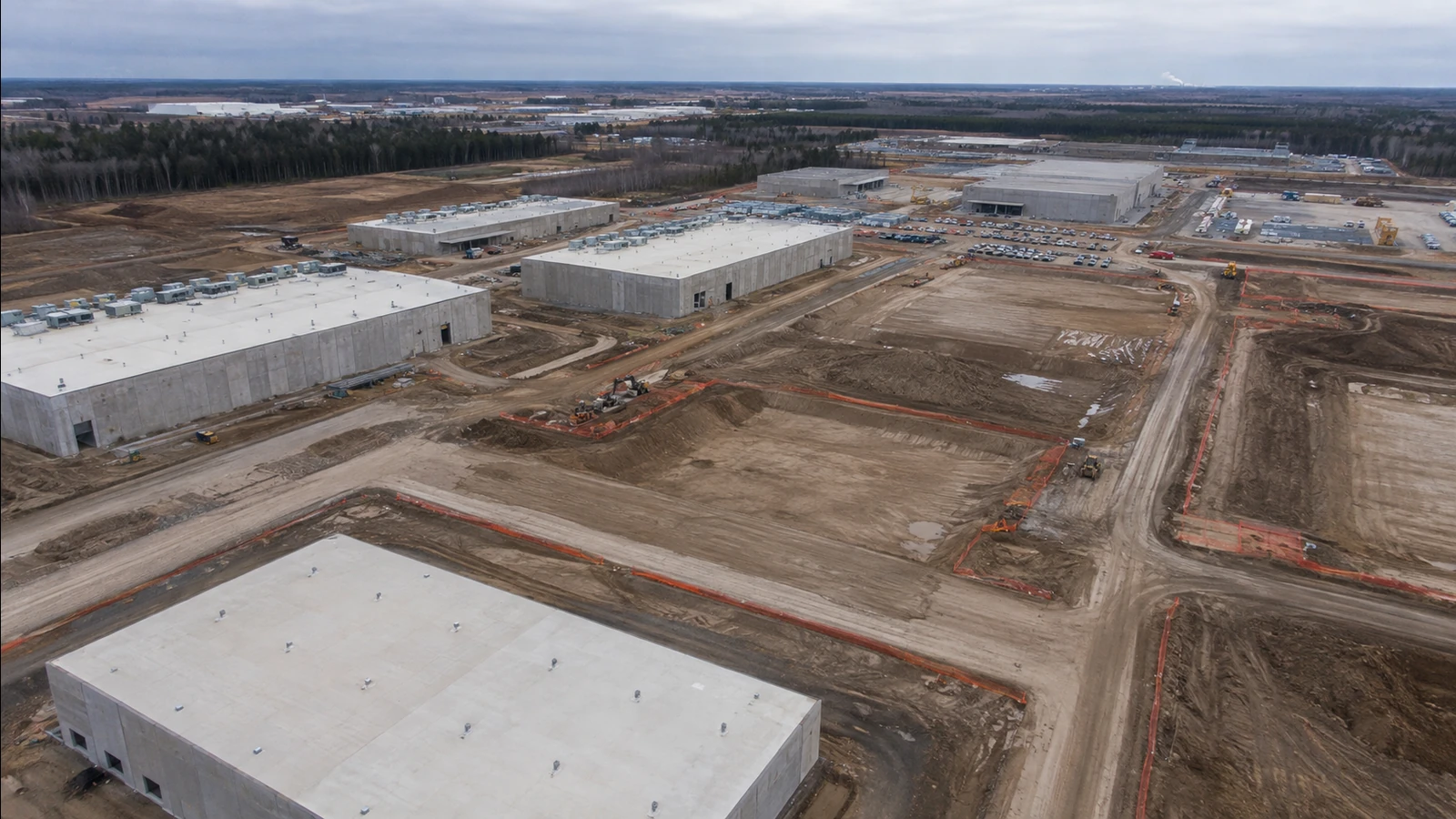 Oblique aerial view of concrete data center buildings and graded construction corridors under an overcast sky