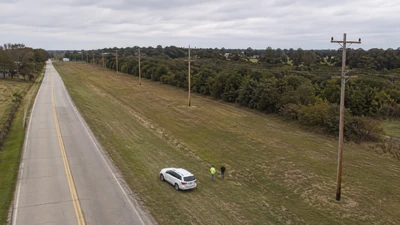 Oblique aerial view of two survey workers beside a white vehicle along a rural road and a line of wooden utility poles