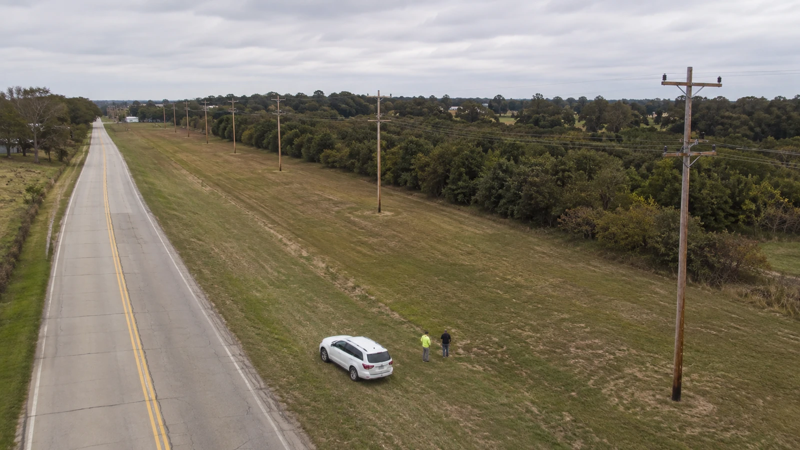 Oblique aerial view of two survey workers beside a white vehicle along a rural road and a line of wooden utility poles