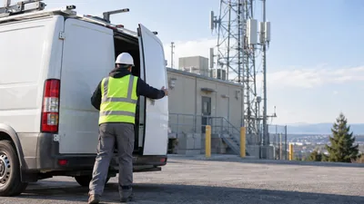 A worker in a hard hat and safety vest opens the rear door of a white cargo van near a hilltop lattice tower and shelter.