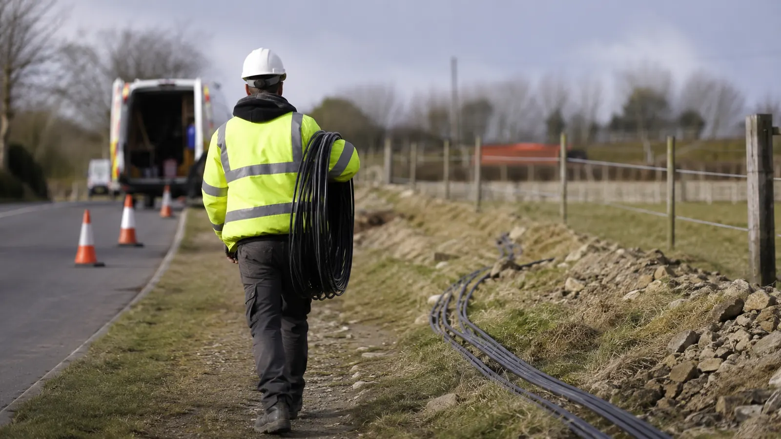 Worker in a hard hat and hi-vis jacket carries a coil of black duct along a coned-off rural roadside.