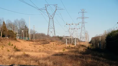 Lattice steel transmission towers and a steel switching structure stand in a dry-grass utility right-of-way, with wooden distribution poles along the tree lines under a clear blue sky.