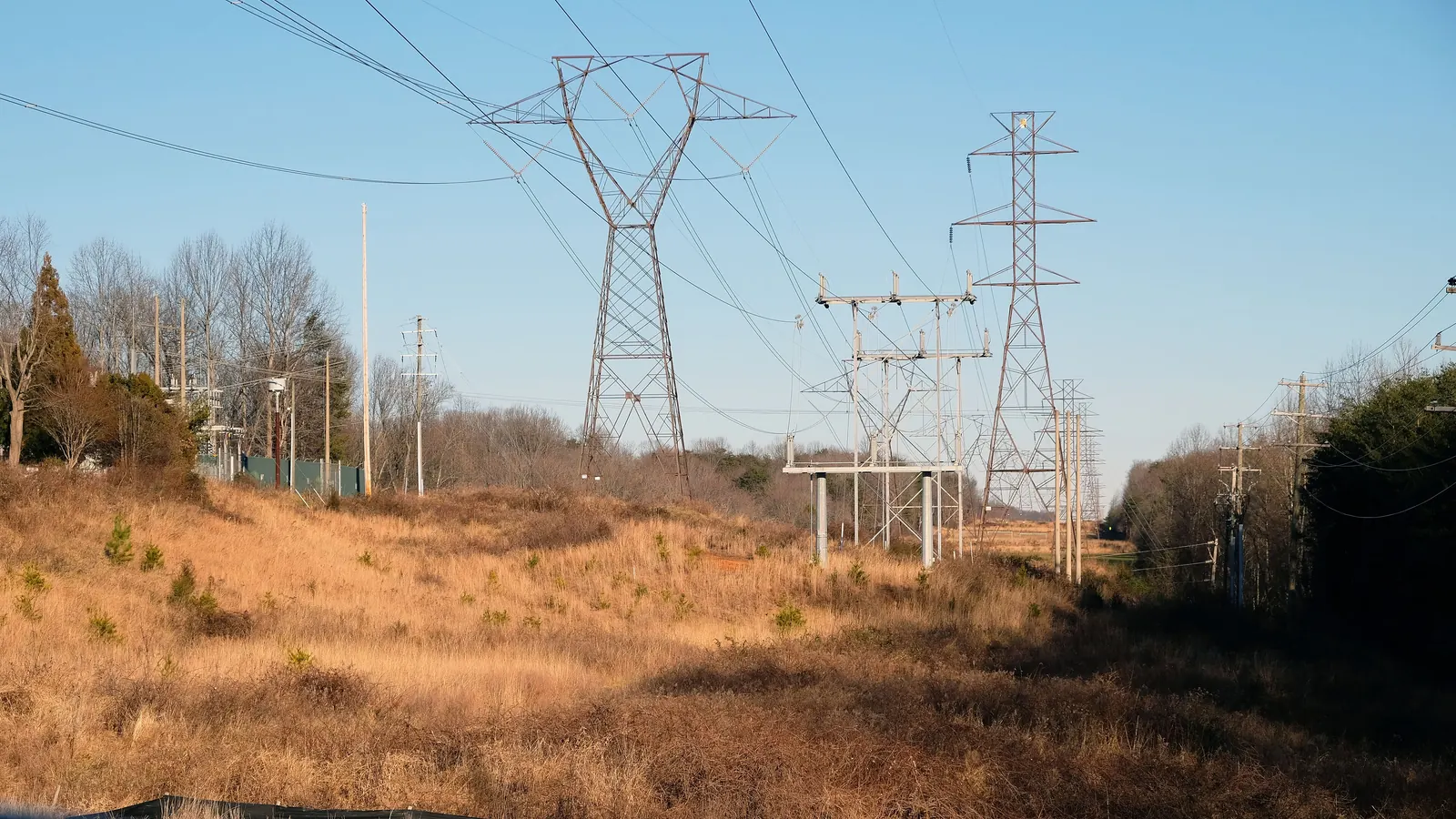 Lattice steel transmission towers and a steel switching structure stand in a dry-grass utility right-of-way, with wooden distribution poles along the tree lines under a clear blue sky.