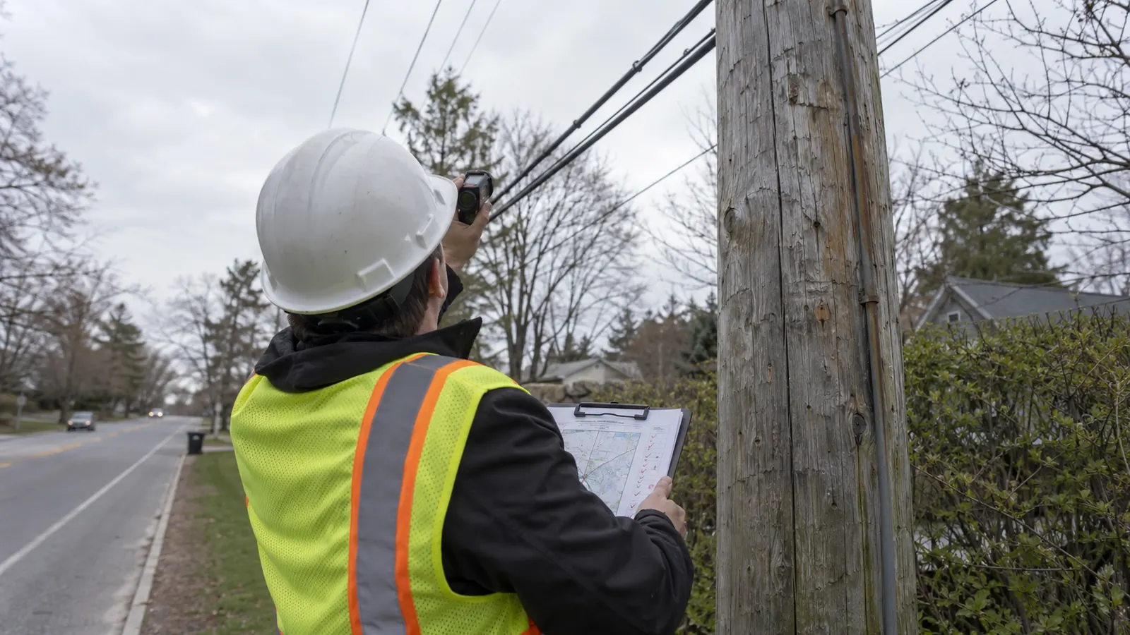 A worker in a hard hat and vest holds a clipboard and aims a handheld device at lines by a wooden roadside pole.
