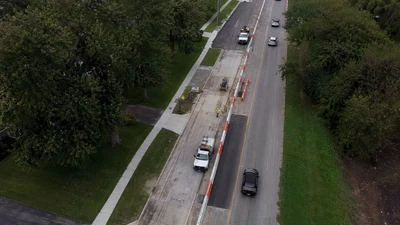 Oblique aerial view of a lane reconstruction zone with temporary barriers, work vehicles, sidewalks, and grass verges
