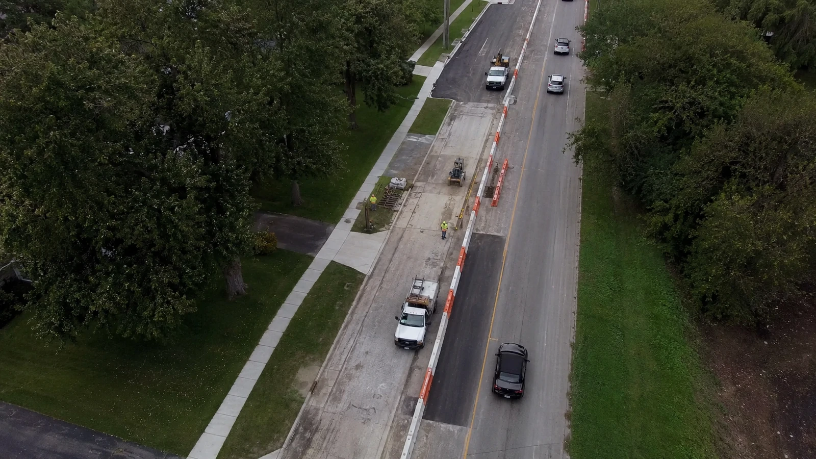 Oblique aerial view of a lane reconstruction zone with temporary barriers, work vehicles, sidewalks, and grass verges