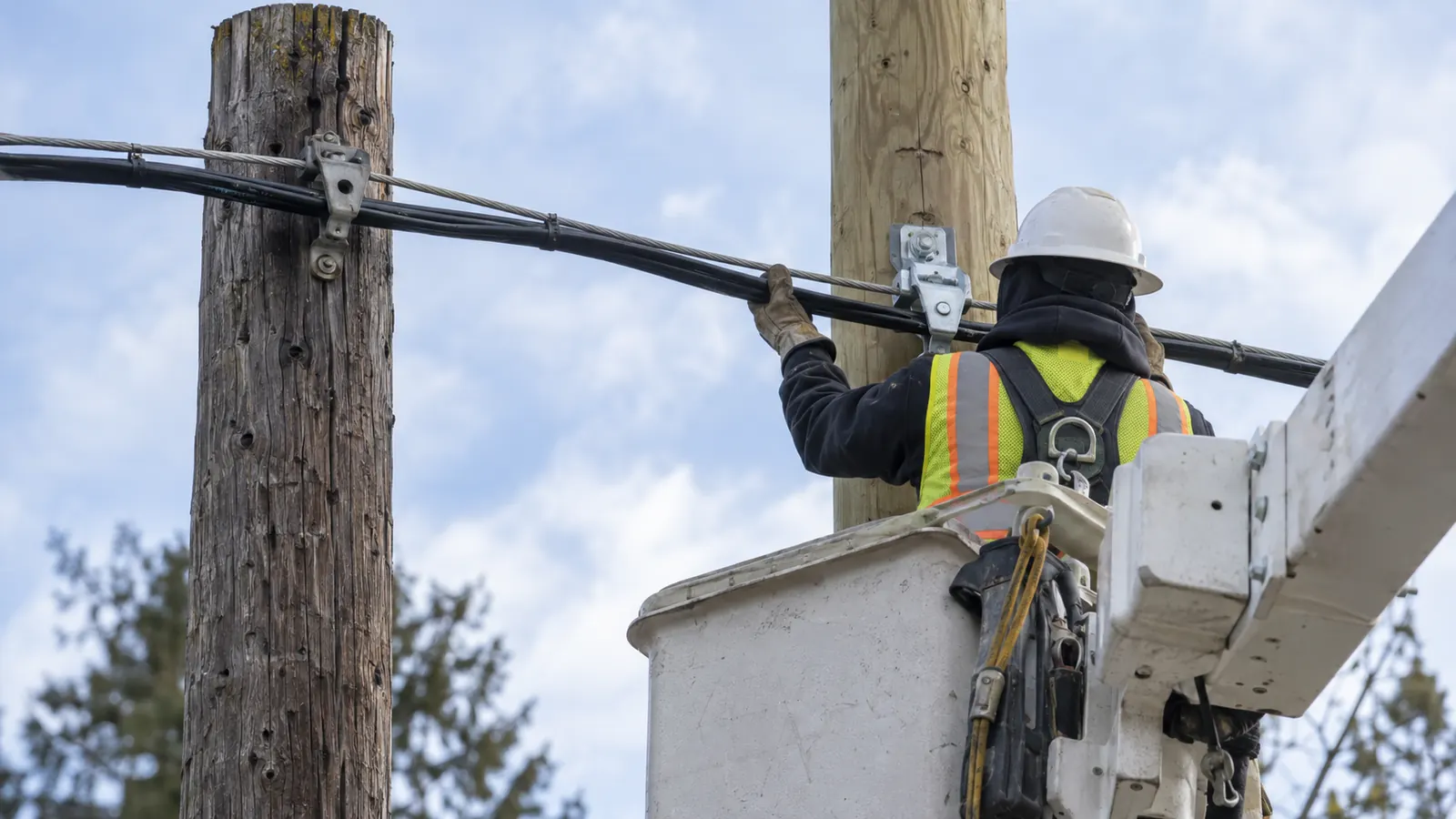 A hard-hatted worker in a white bucket lift grips a cable strung between a weathered wooden pole and a new one.