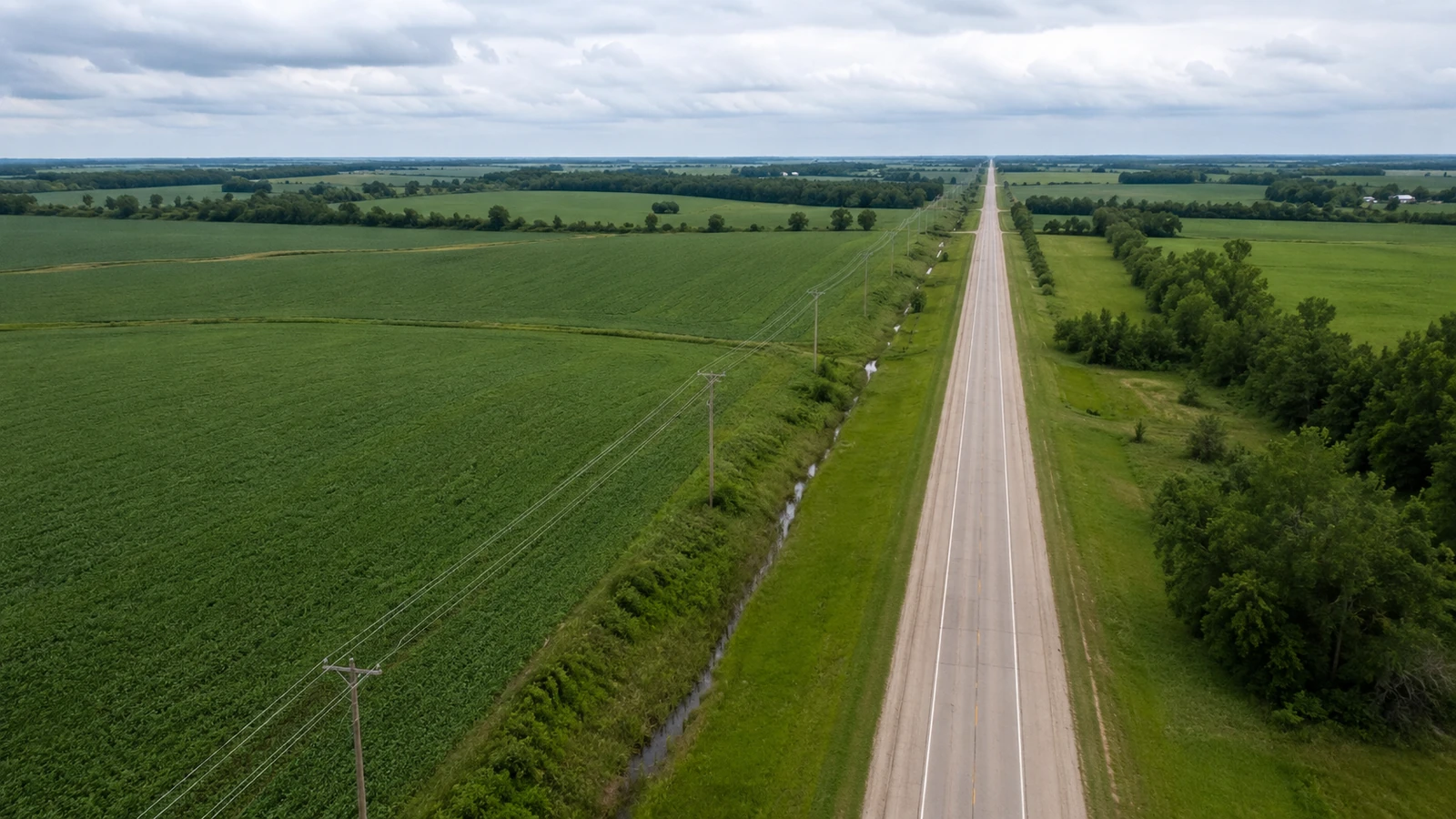 Oblique aerial view of a wooden distribution pole line following a straight rural road between fields and a drainage ditch