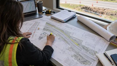 A person in a safety vest marks up a roadway plan sheet at a window desk overlooking a trench and traffic cones.