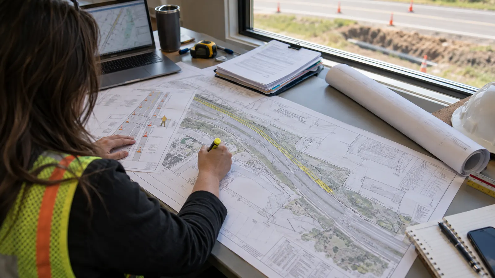 A person in a safety vest marks up a roadway plan sheet at a window desk overlooking a trench and traffic cones.