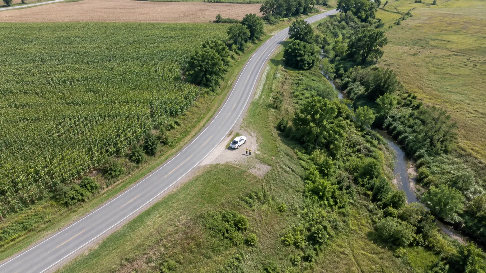 Aerial view of a rural road corridor, fields, drainage, and a parked survey vehicle.