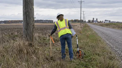 Worker in hard hat and vest sets a flagged stake by a wooden pole beside a gravel farm road, with a measuring wheel.