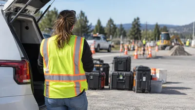 A worker in a safety vest stands at an open white SUV beside equipment cases, facing a coned-off roadside dig with an excavator.