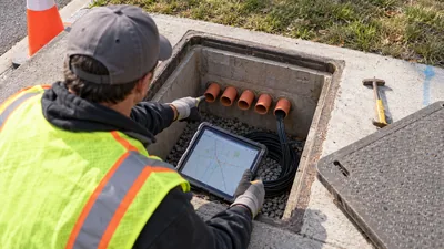 A worker in a safety vest holds a tablet showing a map over an open utility vault and points at its orange conduits.