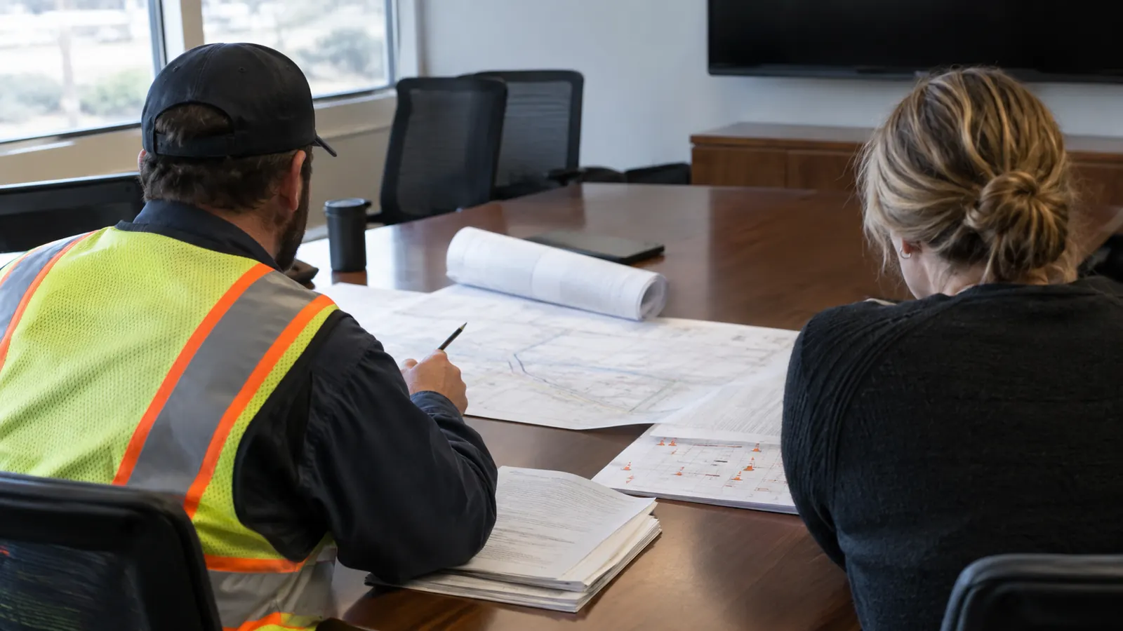 A man in a safety vest holds a pencil over plan sheets as a woman reviews papers at a wooden conference room table.