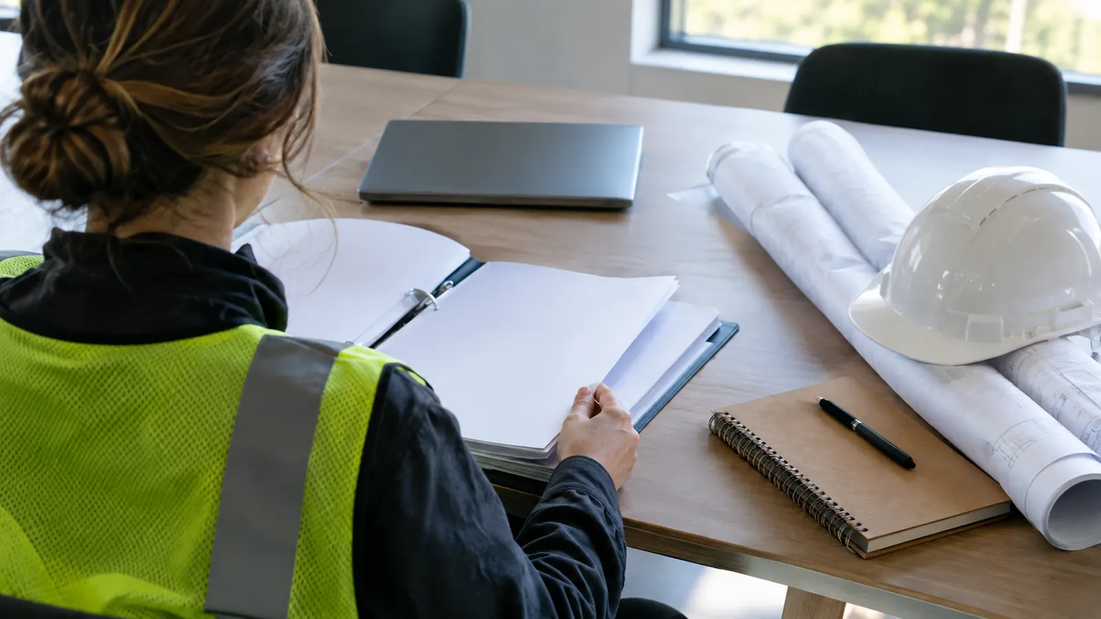 A person in a yellow safety vest reads a binder at an office table beside a laptop, rolled plans and a white hard hat.